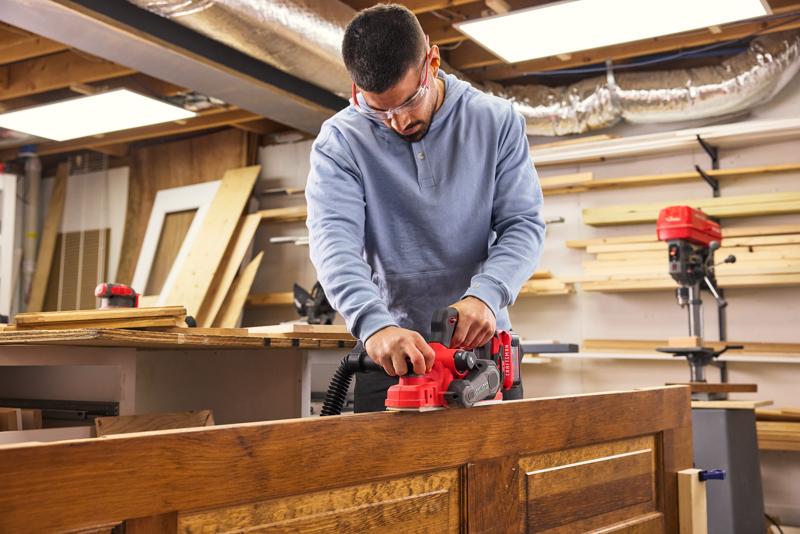Man using CRAFTSMAN V20* Hand Planer on wood door, in wodshop setting.
